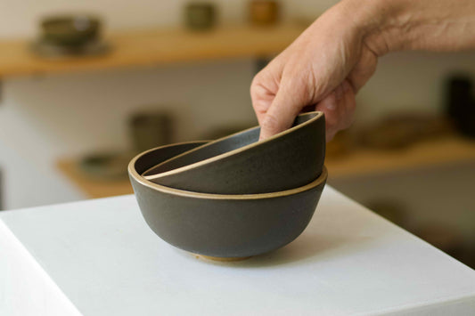 Person lifting a stack of dark ceramic bowls on a light surface.