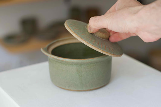 Hand opening a green ceramic lid on a matching pot with a blurred background