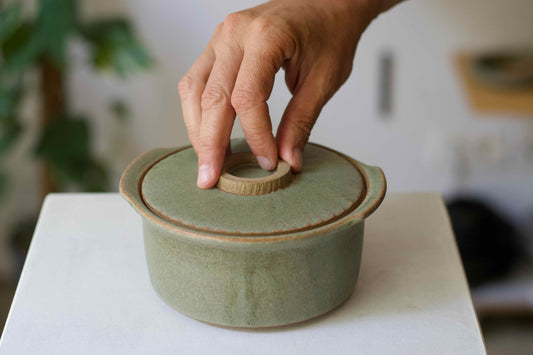 Hand opening a green ceramic lid on a white surface with a blurred background