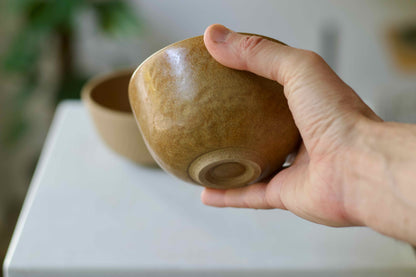 Hand holding a brown ceramic bowl with another bowl in the background on a white surface.